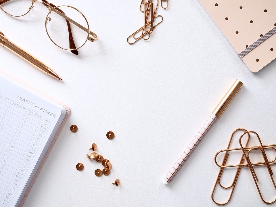 A flat lay photo of rose gold office accessories featuring paper clips, pens, thumbtacks, planners and a pair of eyeglasses